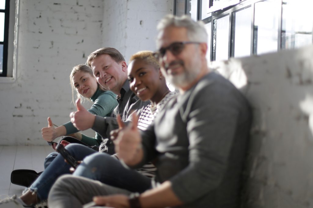 Side view of group of diverse friends of different ages sitting on floor in white room with brick walls and showing thumb up gesture while smiling at camera
